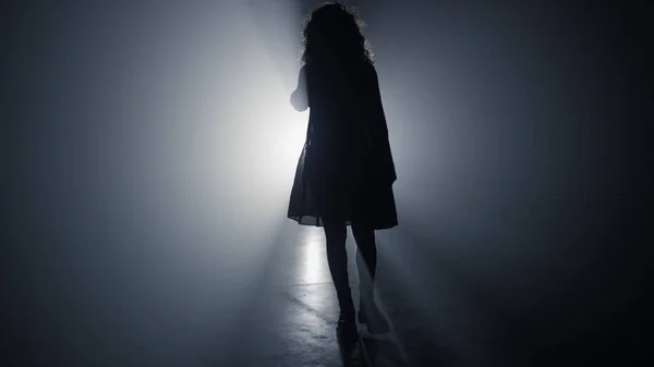 Silhuett kvinna går i mörker. Flicka promenader i strålkastare bakgrund. — Stockfoto