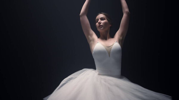 Charming ballet dancer performing classical dance in dark space. Beautiful ballerina moving hands indoors. Slim woman spinning around in tutu skirt on stage.