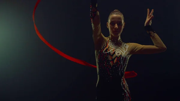 Chica actuando con cinta voladora para cámara. Gimnasta ensayando en salón oscuro. — Foto de Stock