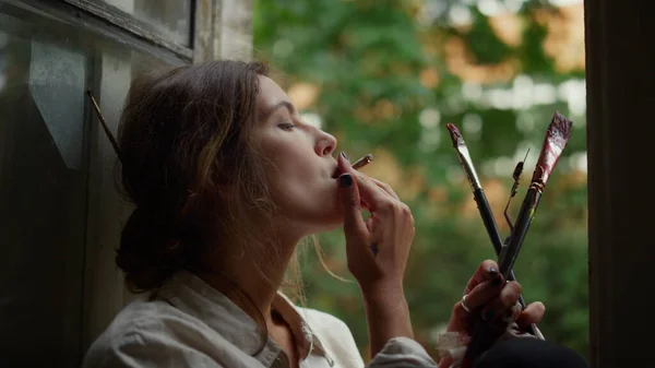Kreativ kvinna som håller cigarett inomhus. Kvinnlig målare röker på fönsterbrädan. — Stockfoto
