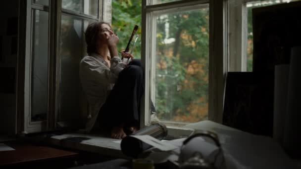 Mulher séria a fumar dentro de casa. Pintor contemporâneo relaxante no peitoril da janela. — Vídeo de Stock