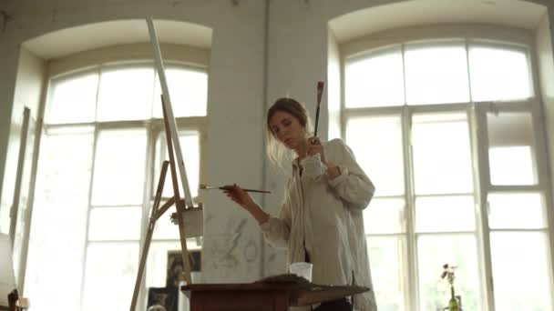 Mujer talentosa sosteniendo cepillos en el interior. Pintor creativo de pie cerca del caballete. — Vídeo de stock