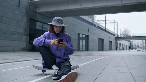 Skater met smartphone outdoor. Hipster zit op skateboard met telefoon. — Stockfoto