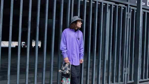 Serieuze kerel poseren in de buurt van stadion poort. Hipster verblijf met skateboard outdoor. — Stockfoto