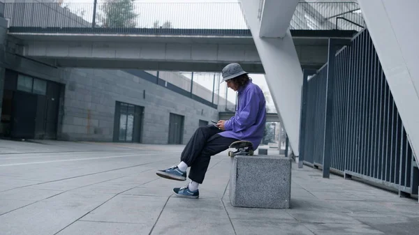 Konzentrierter Mann mit Smartphone im Freien. Junger Hipster sitzt auf Skateboard. — Stockfoto