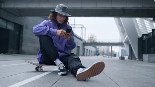 Um hipster desportivo a ler telemóvel no skate. Homem sentado no skate com telefone. — Vídeo de Stock