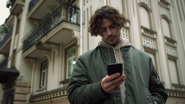 Hombre centrado mensajes de texto en el teléfono inteligente al aire libre. Hipster positivo sosteniendo el teléfono celular. — Foto de Stock