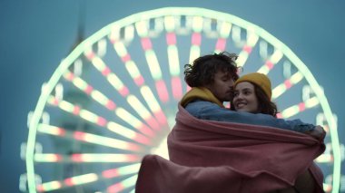 Aşk çifti dönme dolaba karşı mutlu hisseder. Erkek kadını battaniyeye sarar.