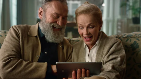 Riendo pareja madura mirando pantalla de la tableta en el sofá en la sala de estar clásica. —  Fotos de Stock