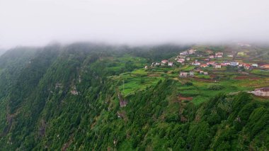 Yeşil tepelerdeki sakin köy huzurlu hava manzarası sunuyor. Manzaralı evler ve gür tarım arazileri sisli gökyüzünün altında güzel dağ yamaçlarına yerleştirildi. Kırsal alan resimsel doğayı vurguluyor 