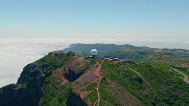 Pico do Arieiro 'daki beyaz küresel meteoroloji istasyonu. Açık mavi gökyüzü altında yeşillikleri kaplayan kayalık uçurumlarla dolu bir hava manzarası. Ünlü yürüyüş rotası başlangıç noktası. Seyahat ve macera.