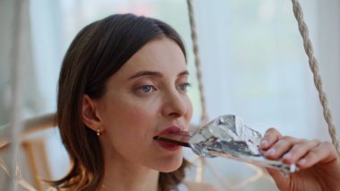 Çikolatayı ısıran güzel bir kadın evde huzur dolu bir memnuniyet gösteriyor. Portre rahatlamış esmer, kahve içen asılmış sandalye. Huzurlu bayan hafif odada içecekle tatlı atıştırmanın tadını çıkarıyor..