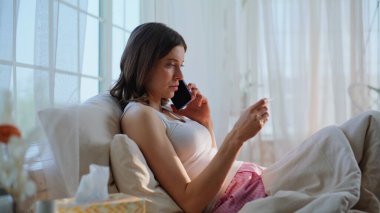 Cep telefonuyla konuşan sağlıksız bir kadın ev yatağında sıcaklığı ölçüyor. Sağlıksız esmer kadın yastıkların üzerinde yatan doktorla telefonda görüşüyor. Hasta kadın akıllı telefon ile hastalıkları tartışıyor..