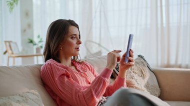 Şaşırmış bir kadın akıllı telefon bankacılığı uygulamasına göz atıyor kanepeye yakın oturuyor. Mutlu genç bayan, internet üzerinden seviniyor. Evdeki koltukta cep telefonuna bakıyor. Heyecanlı duygusal esmer evde harika telefon haberleri okuyor.