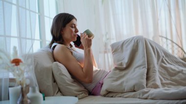 Uyanmış bir kadın rahat bir yatakta sabah kahvesini yudumlarken cep telefonundan konuşuyor. Kaygısız bir kadın cep telefonunu arıyor ve beyaz bir yatağın üzerinde yatıyor. Mutlu esmer konuşan akıllı telefon hafta sonunun keyfini çıkar..