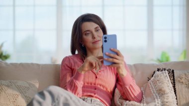 Dinlenen bayan akıllı telefonu izliyor rahat koltukta tek başına oturuyor. Gülümseyen kadın rahat ev koltuğunda rahatlarken cep telefonunu kaydırıyor. Sakin esmer, dairede cep telefonuyla online alışveriş yapıyor..