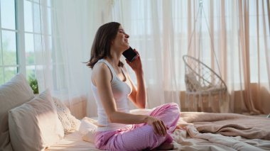 Kadın cep telefonuyla konuşmaya başlıyor. Güneşli yatak odasındaki yumuşak yatağa bağlanmış şekilde oturuyor. Gülümseyen esmer cevaplı akıllı telefon yatakta dinleniyor. Sabahın erken saatlerinde cep telefonuyla konuşan neşeli bir bayan..
