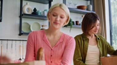 Yakın çekim kankaları mutfak tezgahına eko-kağıt paketleri yerleştiriyor. Marketten gülümseyen kızlar geldi. Alışveriş yapıp evde eğleniyorlardı. Gülen kadınlar birlikte yemek sıralarlar.