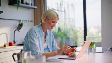Gülümseyen kadın, parlak mutfakta defter başında çalışıyor. Uzaktan kumandalı profesyonel ev ortamında dizüstü bilgisayarı kapatma işi. Rahat bayan serbest çalışan, açık renk saçlı, dinlenen.
