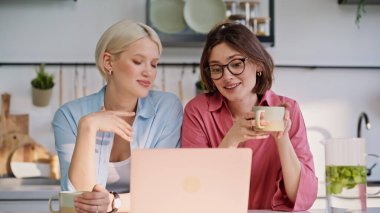 Ev arkadaşları, sakin bir mutfak sabahında bilgisayardan konuşarak ekranı izliyorlar. Gülümseyen lezbiyen çift, mutfakta kahve içip laptoplarını arıyor. Mutlu kadınlar internetten konuşan içeceklerden hoşlanır.