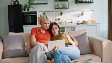 Evde konuşan tablet kullanan kızları kucaklamak. Neşeli Igbt çifti oturma odasında modern ped bilgisayarlarını tutarak kanepeye sarılıyorlar. Pozitif kız arkadaşlar içeride eğlenceli videolar izlemekten hoşlanırlar.