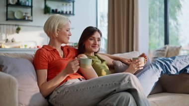 Kanepede çay fincanı tutan lezbiyenleri kucaklamak. Igbt çiftlerinin evde birlikte kahve içmelerini seviyorum. Gülümseyen kız arkadaşlar sıcak içecek içiyorlar rahatlatıcı kanepe. İnsanların yaşam tarzı 