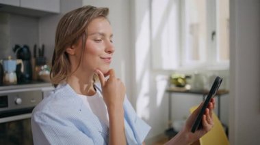 Güneşli mutfakta neşeli bayan mesajlaşması. Kapalı mekanda cep telefonu kullanan bir kadın. Pijamalı gülümseyen bir kadın sosyal medyada geziniyor. Pozitif genç model teknolojiyle etkileşimde.
