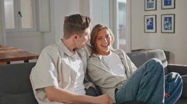 Ev sahibi çift gün ışığında sohbet ediyorlar. Yakın partnerler kanepede birbirlerine sarılıp konuşuyorlar. Gülümseyen adam utangaç kız arkadaşını kucaklıyor ve evde rahat bir hafta sonu geçiriyor. Romantik bağlantı kavramı.