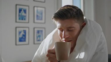 Adam battaniyeye sarılı ev kahvesi içiyor. Portre, sabah güneşinde sıcak bir içecek içip bilinmeyen partnerle konuşmaktan zevk alıyor. Sıcak atmosferde kahvaltı yapan gülümseyen yakışıklı erkek. Ev eğlencesi yaşam tarzı
