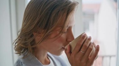 Ev penceresinin yanında kahvesini yudumlayan yakın plan kadın. Tatminkar bayan içeceği tadıyor. İçeride sakin bir sabah atmosferinin tadını çıkarıyor. Huzurlu gülümseyen kız kameraya bakıyor.. 