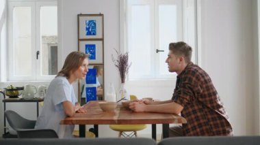 Sakin bir sabahta mutfak masasıyla iletişim kuran rahat ortaklar. Gülümseyen çift kahvaltısı modern iç mekanda konuşuyor. Genç bir kadın hafta sonu yemeğinden zevk alıyor. Mutlu eşler yaşam tarzı kavramı.