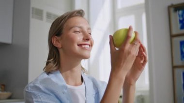Güneşli mutfakta lezzetli elmaları ısıran bir kadın. Kapanış kızı taze meyvelere hayran evde huzurlu bir sabah geçiriyor. Sağlıklı atıştırmalıklar yiyen gülümseyen kadın içeride kameraya bakıyor. Refah diyeti konsepti.