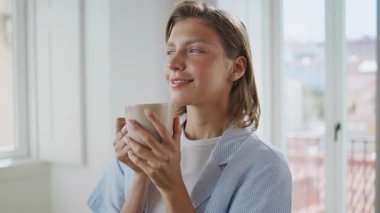 Serene Lady sabah evinde bardağı tutuyor. Yakın plan. Gülümseyen kız huzurlu bir hafta sonunda kahve içmenin tadını çıkarıyor. Pijamalı, gözleri kapalı mutlu bir manken. Rahat bir yaşam tarzı reklamı.