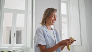 Mutfakta muz soyan neşeli kadın kendini mutlu hissediyor. Mutlu kız kahvaltıda sağlıklı abur cubur diyetinin tadını çıkarıyor. Gevşemiş kısa saçlı öğrenci iç mekanda meyve tutuyor. İnsan yaşam tarzı.