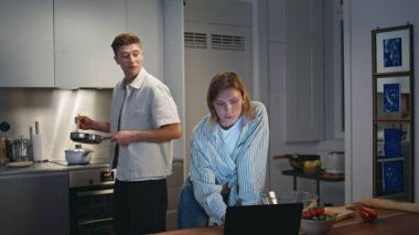 Modern bir çift evde yemek hazırlıyor. Gülümseyen eş ev yemeği tadıyor, neşeli kocalar konuşuyor. Genç aile hafta sonları birlikte akşam yemeği pişiriyor. Mutfakta bilgisayarla çalışan bir kadın.