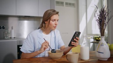 Modern mutfakta mesajlaşan kadınları yemek. Yakın çekim. Masada cep telefonu mesajı okuyan gülümseyen kız. Rahatlamış bir kadın evde sabah rutininin tadını çıkarıyor. Modern aygıt karıştırma kasesi kullanarak günlük model