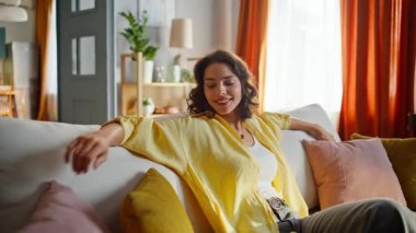 Oturma odasında rahat rahat gülümseyen bayan kanepe yastıklarına dokunuyor. Mutlu kadın rahat koltuğunda oturmuş, modern apartmanın tadını çıkarıyor. Kaygısız kıvırcık esmer yalnız başına takılıyor. Yurt içi dinlenme