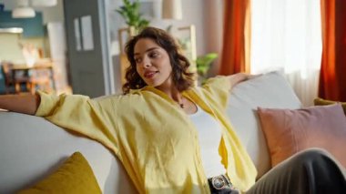 Carefree girl falling sofa looking around modern living room closeup. Portrait smiling woman sitting comfortable couch at calm domestic weekend. Happy woman wearing yellow shirt chilling in cozy home.