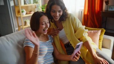 Besties video araması cep telefonu oturma odasındaki kanepede rahat bir şekilde oturuyor. Gülümseyen lezbiyen çift dairede cep telefonu webcam ile konuşuyor. Kanepede akıllı telefon görüşmesinin keyfini çıkaran iki pozitif arkadaş..