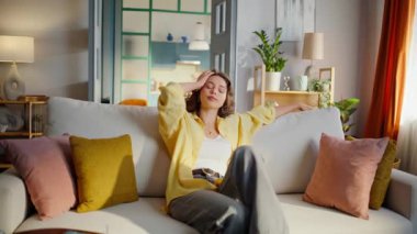 Sarı gömlek giyen yorgun ve rahat bir kadın. Huzurlu genç bayan rahat bir kanepede oturuyor ve evin iç kısmının tadını çıkarıyor. Kıvırcık esmer sakin bir ev ortamında dinleniyor..