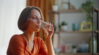 Esmer kadın, modern mutfak iç mekanında tek başına oturmuş tatlı su içiyor. Güzel, genç bir kadın camdan taze bir içecek yudumlarken gülümsüyor. Mutlu bayan sabah ferahlatıcı. Sağlıklı beslenme.