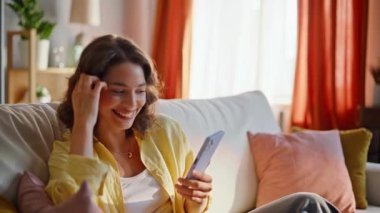 Happy student reading message mobile phone relaxing on cozy home couch closeup. Smiling woman browsing cellphone sms sitting comfortable sofa. Relaxed lady watching smartphone content in light room.