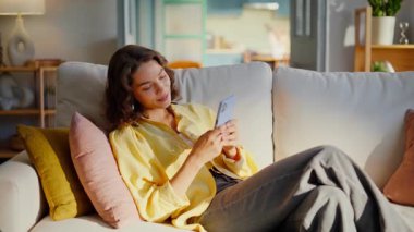 Joyful brunette texting mobile phone enjoying domestic leisure on comfortable sofa closeup. Smiling woman typing cellphone message sitting couch. Happy student shopping online using smartphone app.