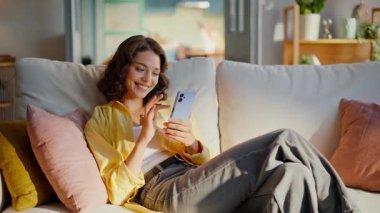 Neşeli kız cep telefonuyla mesajlaşıyor yumuşak kanepe yastıklarının üzerinde rahatça oturuyor. Gülümseyen kadın akıllı telefon uygulaması kullanarak internet alımı yapıyor. Mutlu esmer evde internet iletişiminin keyfini çıkarın..