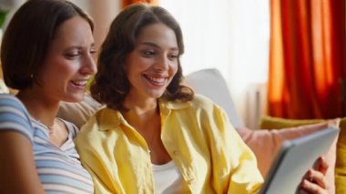 Tablete bakan birkaç kadın evdeki kanepede online alışverişi tartışıyor. İki kız arkadaş bilgisayarda ekommerce uygulamasına göz atıyorlar. Rahat kanepede oturan rahat kankalar.