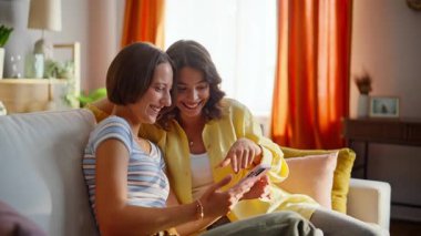 Joyful besties looking smartphone relaxing on comfortable apartment sofa closeup. Happy two friends laughing browsing cellphone content on couch. Smiling love couple reading funny message together.