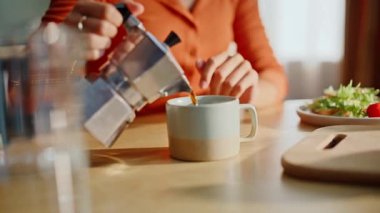 Kadın elleri, ahşap mutfak masasındaki kahve makinesinden kahve dolduruyor. Evde rahatlamış bir bayan bardağı taze kafein içeceğiyle dolduruyor. Tek başına sabah içkisini yudumlayan pozitif kız..