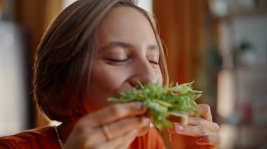 Yakın plan kadın sandviçi ısırıyor ve evde lezzetli yemeklerin tadını çıkarıyor. Sabahleyin yemek yiyen genç bayanın portresi. Salata yapraklı güzel esmer çiğneme yemeği sağlıklı beslenmenin tadını çıkarıyor..