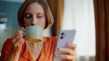 Closeup lady drinking tea reading cellphone sms in apartment kitchen. Portrait beautiful young woman sipping fresh coffee watching smartphone. Smiling brunette enjoying tasty morning beverage at home