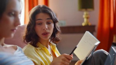 İki arkadaş ev ödevi yapıyor. Yakın çekim. Kıvırcık esmer, elinde kitapla ev zemininde oturan en iyi bilgisayarla konuşuyor. İnternette bilgisayar okuyan rahat öğrenciler..